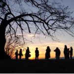 Einige Menschen stehen im Kreis neben einer Linde vor einem Sonnenuntergang in Gemeinschaft
